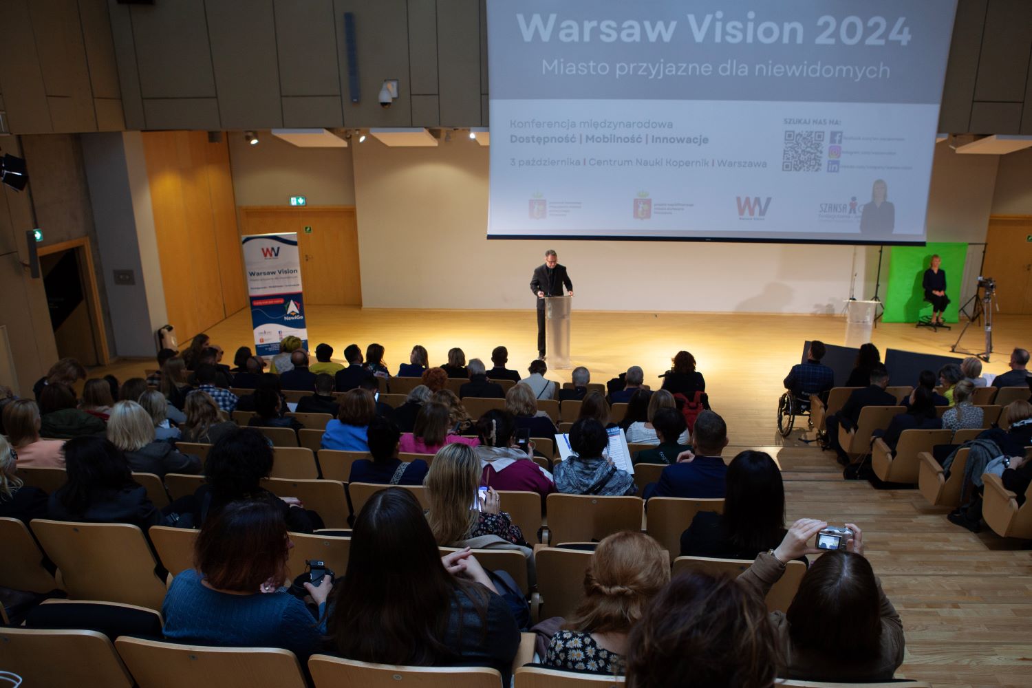 Sala audytoryjna w Centrum Nauki Kopernik. Widok z góry na scenę. Sala wypełniona ludźmi siedzącymi na krzesłach. Na scenie prowadzący Maciej Iwański. Na ekranie z rzutnika  plansza witająca na konferencji Warsaw Vision.