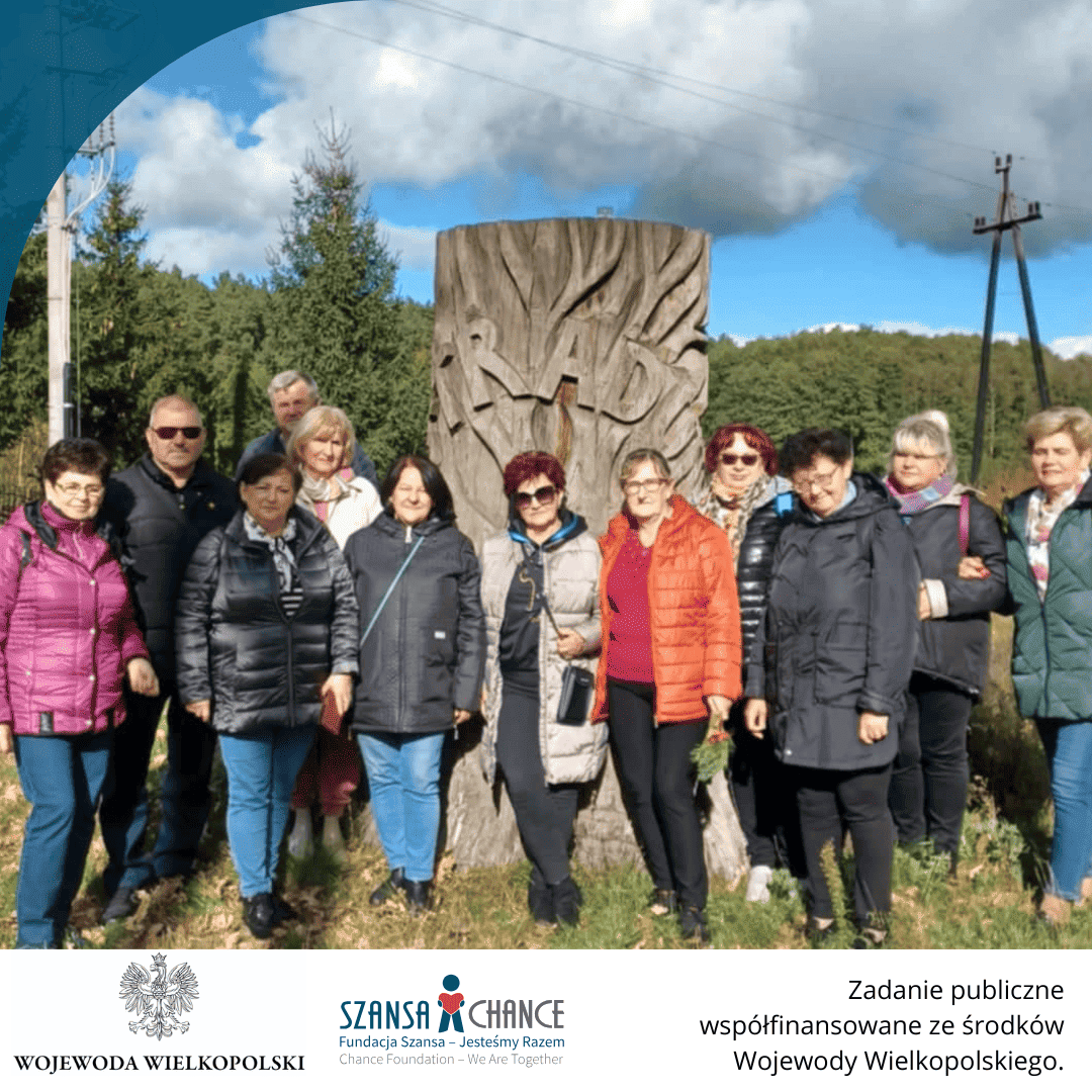 Grupa osób pozuje przy drewnianej rzeźbie na skraju lasu. Osoby są ubrane w kurtki, co wskazuje na chłodniejszą pogodę. Wszyscy uśmiechają się do zdjęcia, tworząc atmosferę wspólnego wypoczynku. Na dole ponownie widnieją loga Wojewody Wielkopolskiego oraz Fundacji Szansa, a także informacja o współfinansowaniu projektu.